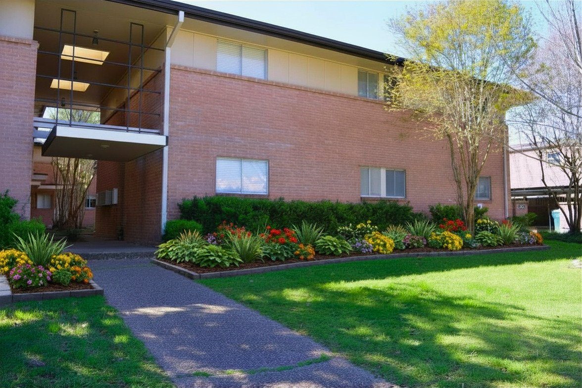 Two-story brick apartment building with landscaped garden beds along the walkway.